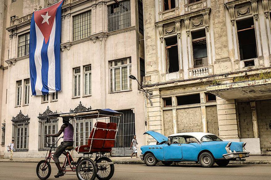 Homem passa de bicicleta em frente a um carro vintage diante de um edifício com uma bandeira de Cuba, nesta terça-feira (31), em Havana