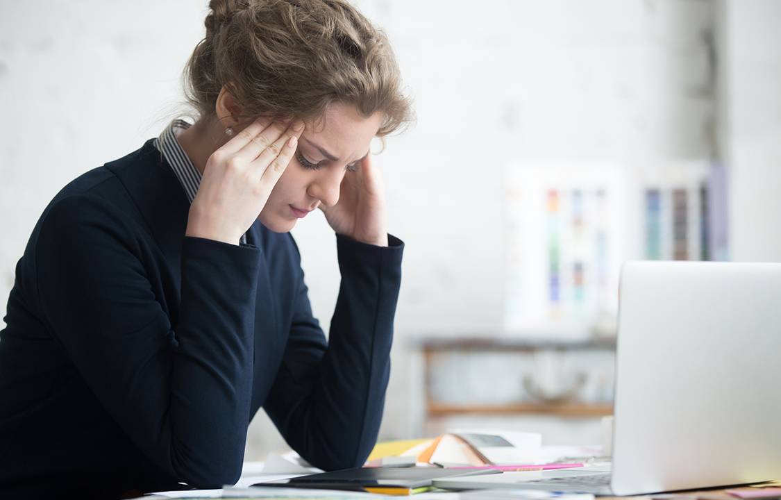 Portrait of young stressed woman sitting at home office desk in front of laptop, touching head with frustrated facial expression, having headache, overworked or depressed