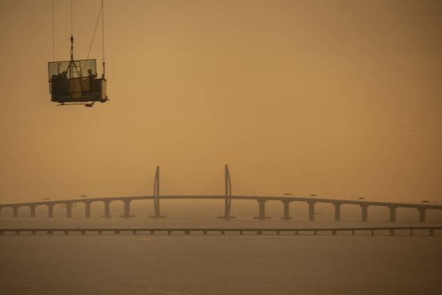 Trabalhadores carregam um painel de vidro em uma plataforma móvel elevada com um trecho da ponte Hong Kong-Zhuhai-Macau ao fundo em Zhuhai, na China - 22/10/2018 Trabalhadores carregam um painel de vidro em uma plataforma móvel elevada com um trecho da ponte Hong Kong-Zhuhai-Macau ao fundo em Zhuhai, na China - 22/10/2018