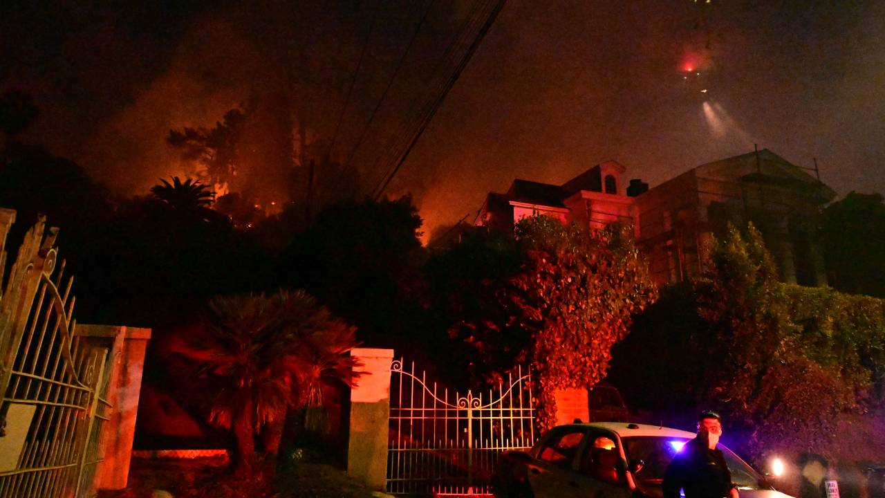 A police officer stands below burning brush at the Sunset Fire near Hollywood Blvd in the Hollywood Hills in Los Angeles, California, on January 8, 2025. At least five people have been killed in wildfires rampaging around Los Angeles, officials said on January 8, with firefighters overwhelmed by the speed and ferocity of multiple blazes -- including in Hollywood. (Photo by Robyn Beck / AFP)