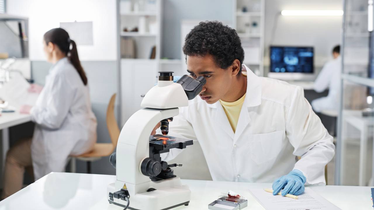 Portrait of young ethnic man as scientist looking in microscope while doing research in laboratory, copy space