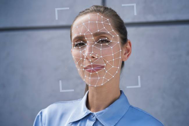 Como funciona reconhecimento facial biometria Mulher branca parada em frente a uma parede. Em seu rosto, projeções computacionais mostram como age o sistema de reconhecimento facial