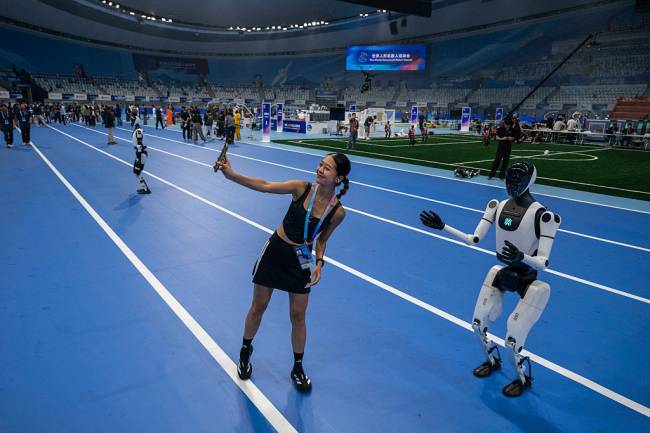 2025 World Humanoid Robot Games – Opening Ceremony BEIJING, CHINA - AUGUST 14: A woman poses with a humanoid robot on the track at the opening ceremony of the World Humanoid Robot Games at the National Speed Skating Oval on August 14, 2025 in Beijing, China. China is holding the event from August 14-17, billed as the first of its kind. (Photo by Kevin Frayer/Getty Images)