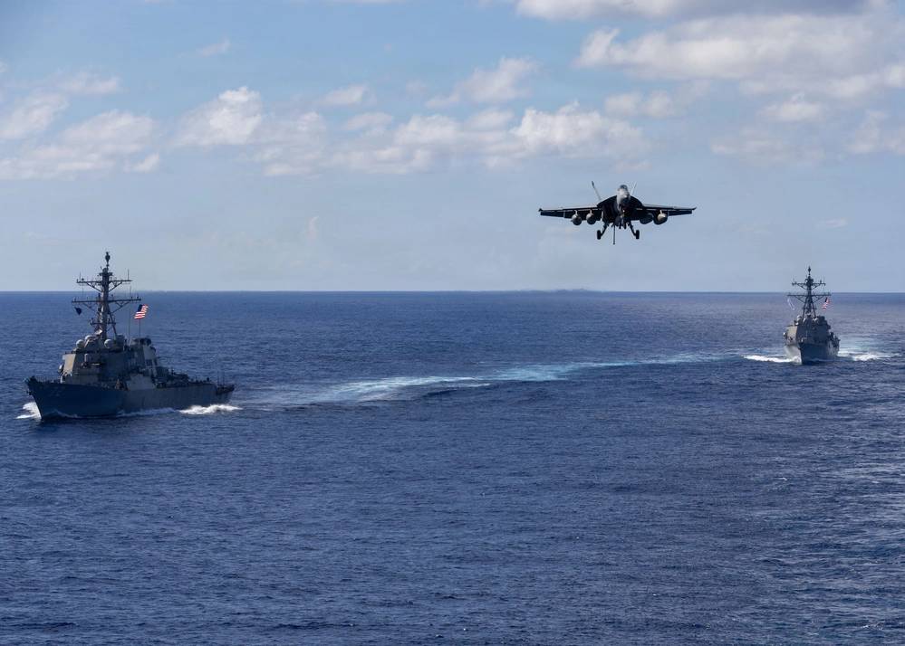 Os destróieres de mísseis guiados da classe Arleigh Burke, USS Mahan (DDG 72), à esquerda, e USS Bainbridge (DDG 96), navegam em formação enquanto um F/A-18 Super Hornet, pertencente à Ala Aérea Embarcada Oito, a bordo do USS Gerald R. Ford (CVN 78), inicia a aproximação final para pouso no porta-aviões. 13/11/2025