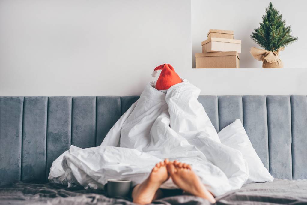 A woman is sitting up in bed, wrapped completely in a white duvet and wearing a Santa hat with a moody, bored expression.