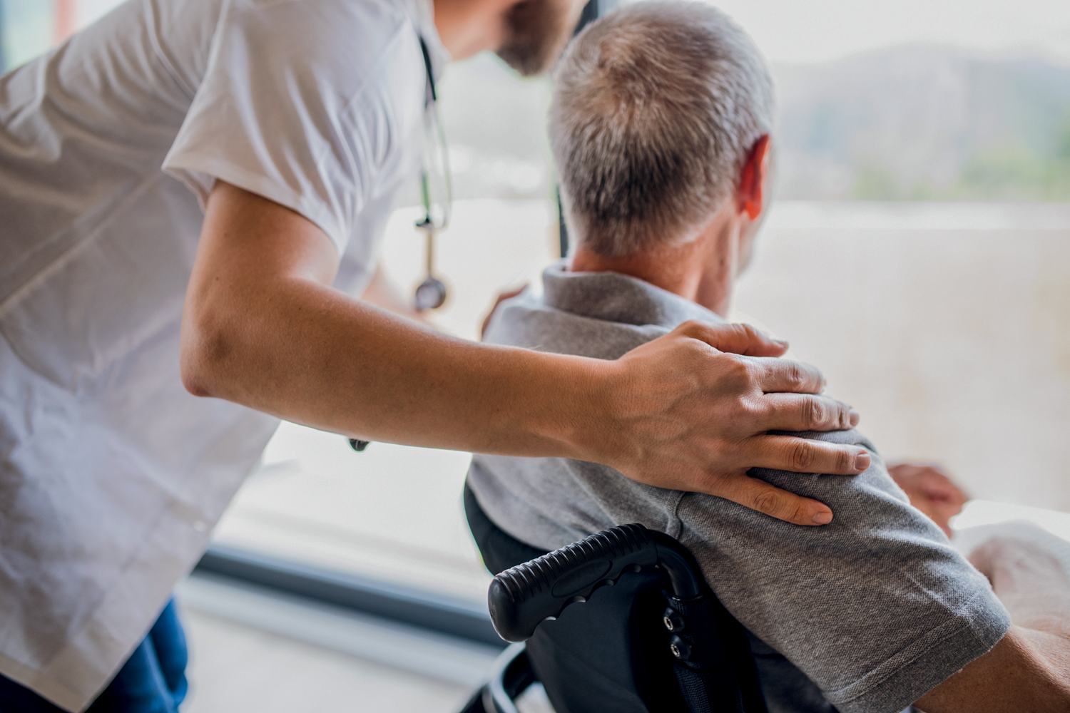 A mature man caregiver with stethoscope and old patient looking out through window. Credito: Halfpoint Images/Getty Images