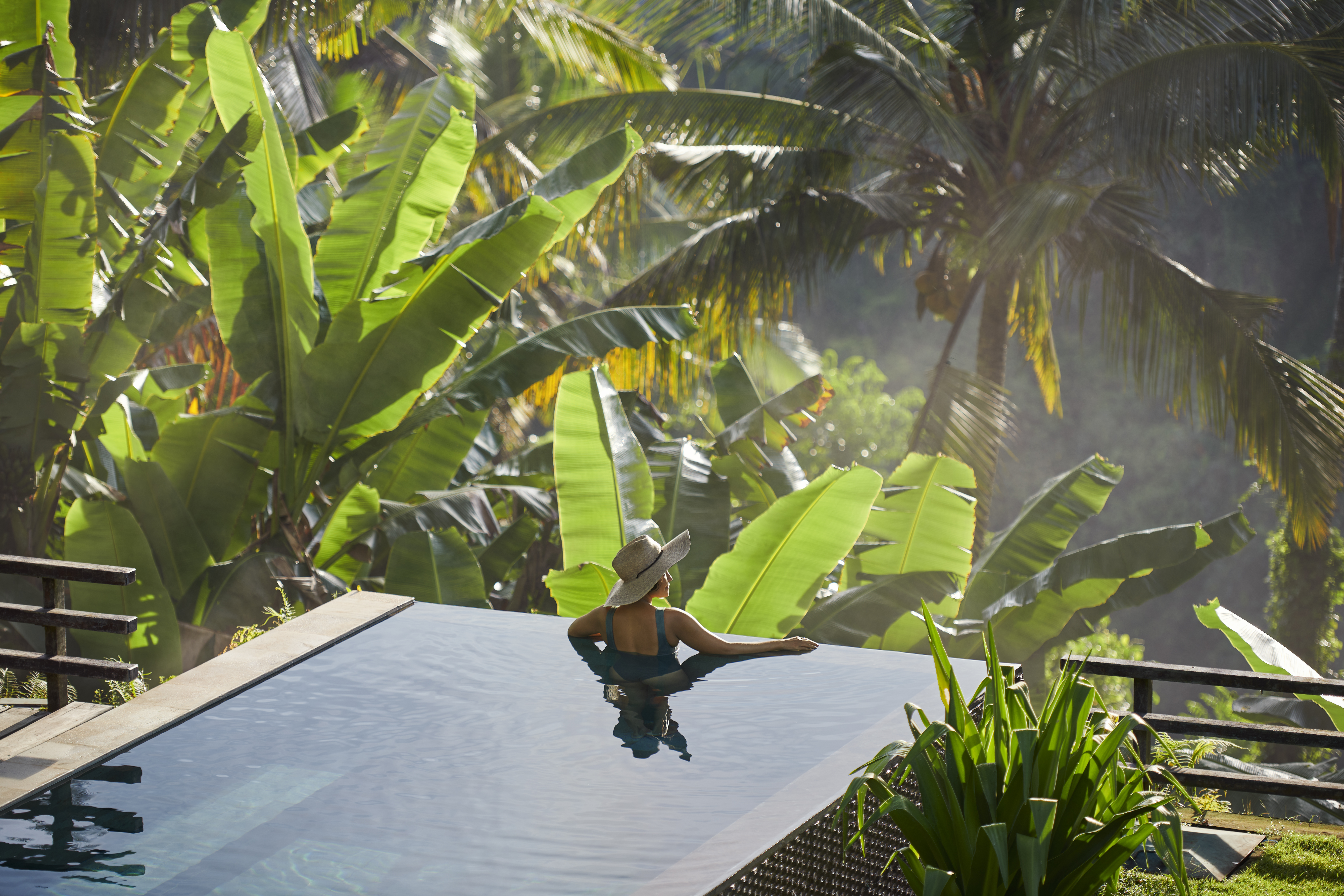 Mature fit Asian woman relaxing on edge of infinity pool, admiring jungle view with early morning mist