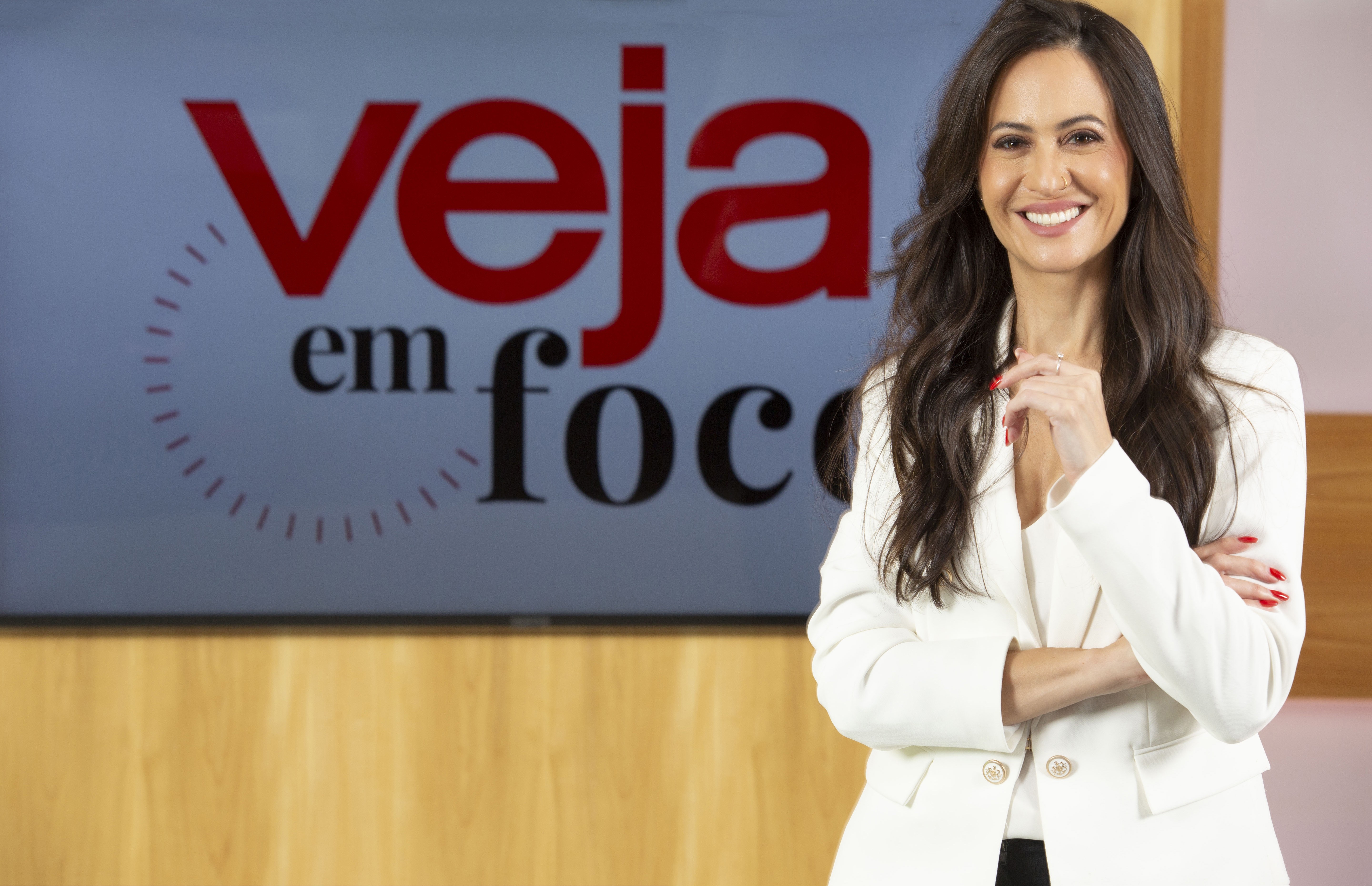 Mulher de cabelos longos e escuros, sorrindo, vestindo blazer branco e blusa decotada, com as mãos cruzadas e unhas vermelhas, em frente a um painel com a logo "Veja em Foco" em vermelho e preto