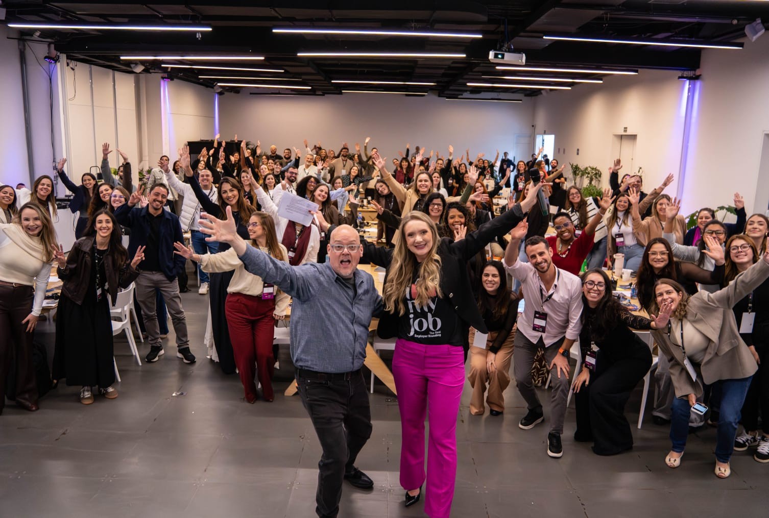 Grande grupo de pessoas diversas, sorrindo e acenando com as mãos para a câmera em um evento ou conferência. Um homem de óculos e camisa azul e uma mulher de calça rosa e blusa preta com a frase "my job" estão em primeiro plano, ambos sorrindo e acenando. O ambiente é um salão amplo com iluminação moderna e mesas ao fundo.