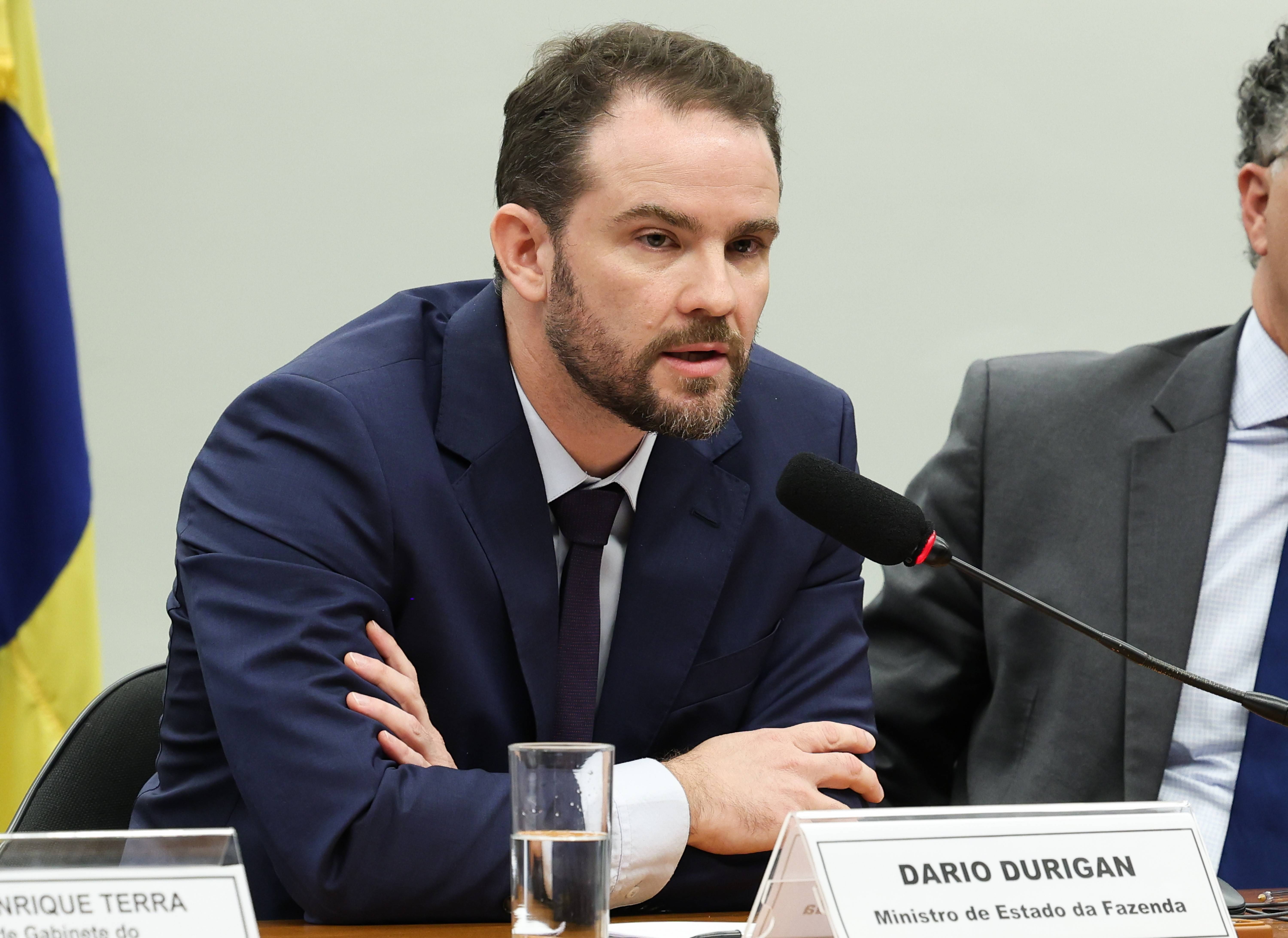 Homem branco de barba, cabelo castanho, terno azul-marinho e gravata escura, falando ao microfone em uma mesa de reunião. À frente, um copo d'água e uma placa de identificação com "DARIO DURIGAN Ministro de Estado da Fazenda".