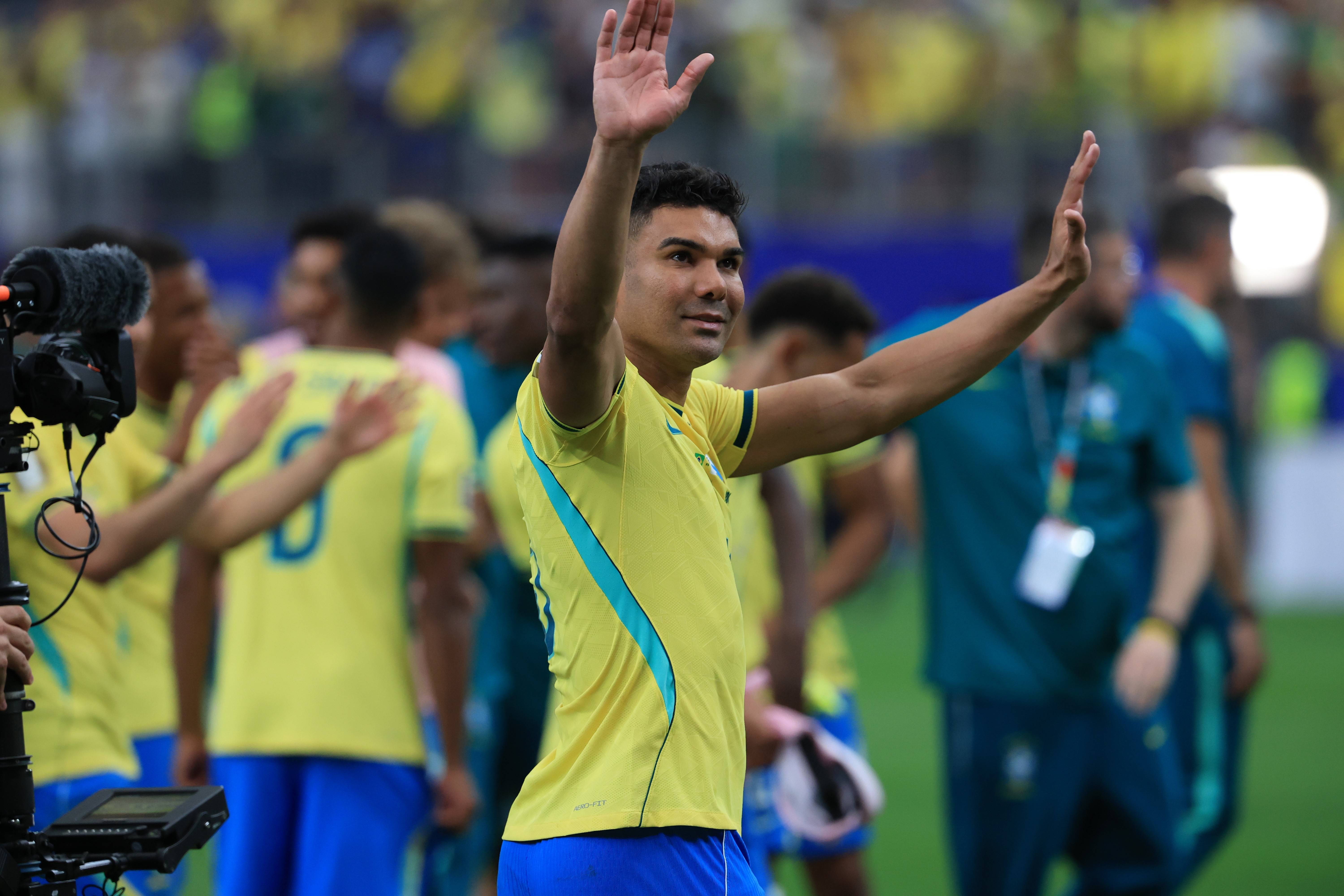 Jogador de futebol Casemiro, com camisa amarela e detalhes azuis, sorri e acena com as duas mãos para a torcida em campo, com outros jogadores e equipe técnica ao fundo