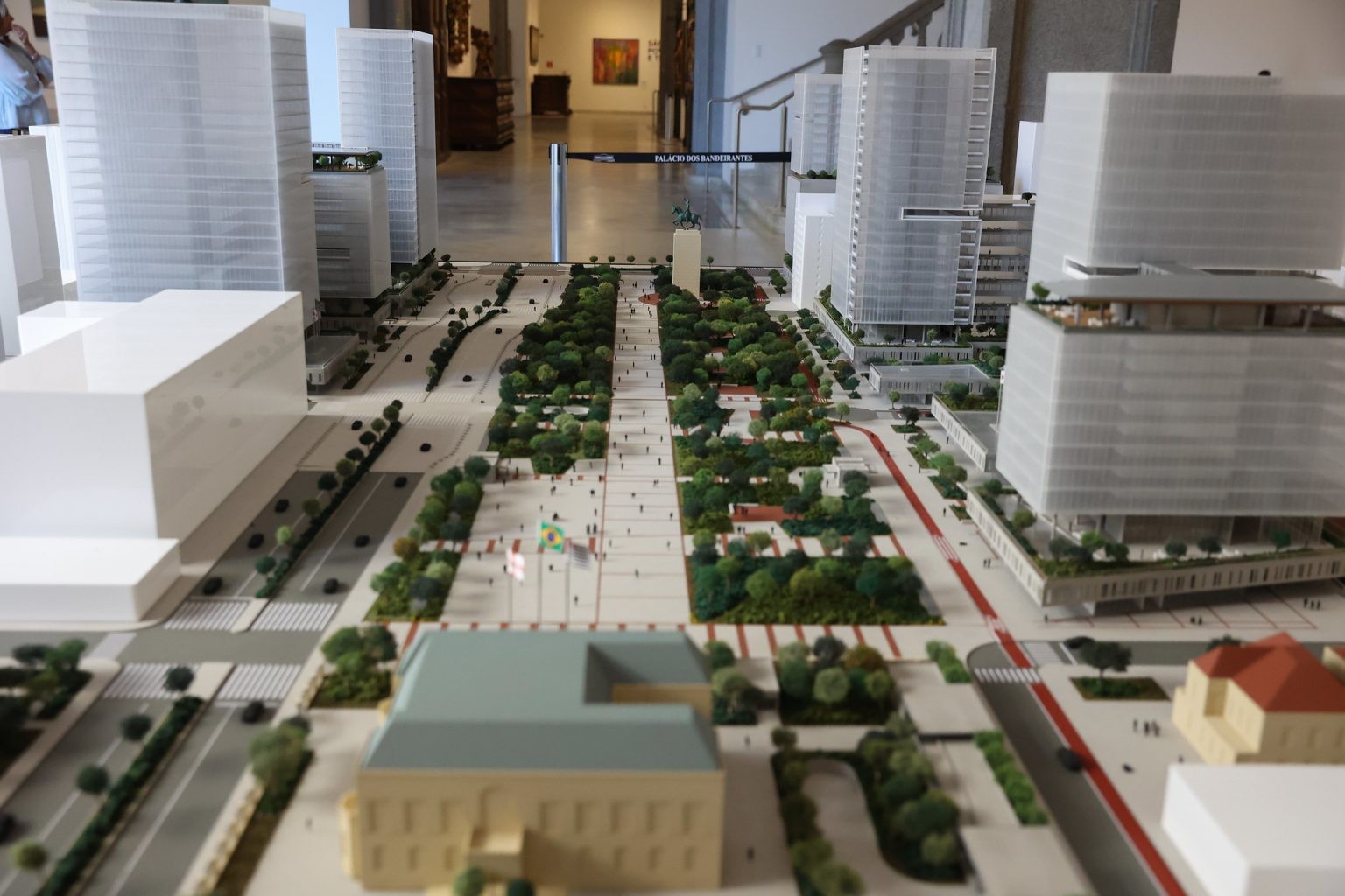 Maquete de uma cidade moderna com arranha-c&eacute;us brancos, avenidas arborizadas, pra&ccedil;as com pessoas em miniatura e edif&iacute;cios baixos, exibida em um ambiente interno com obras de arte ao fundo