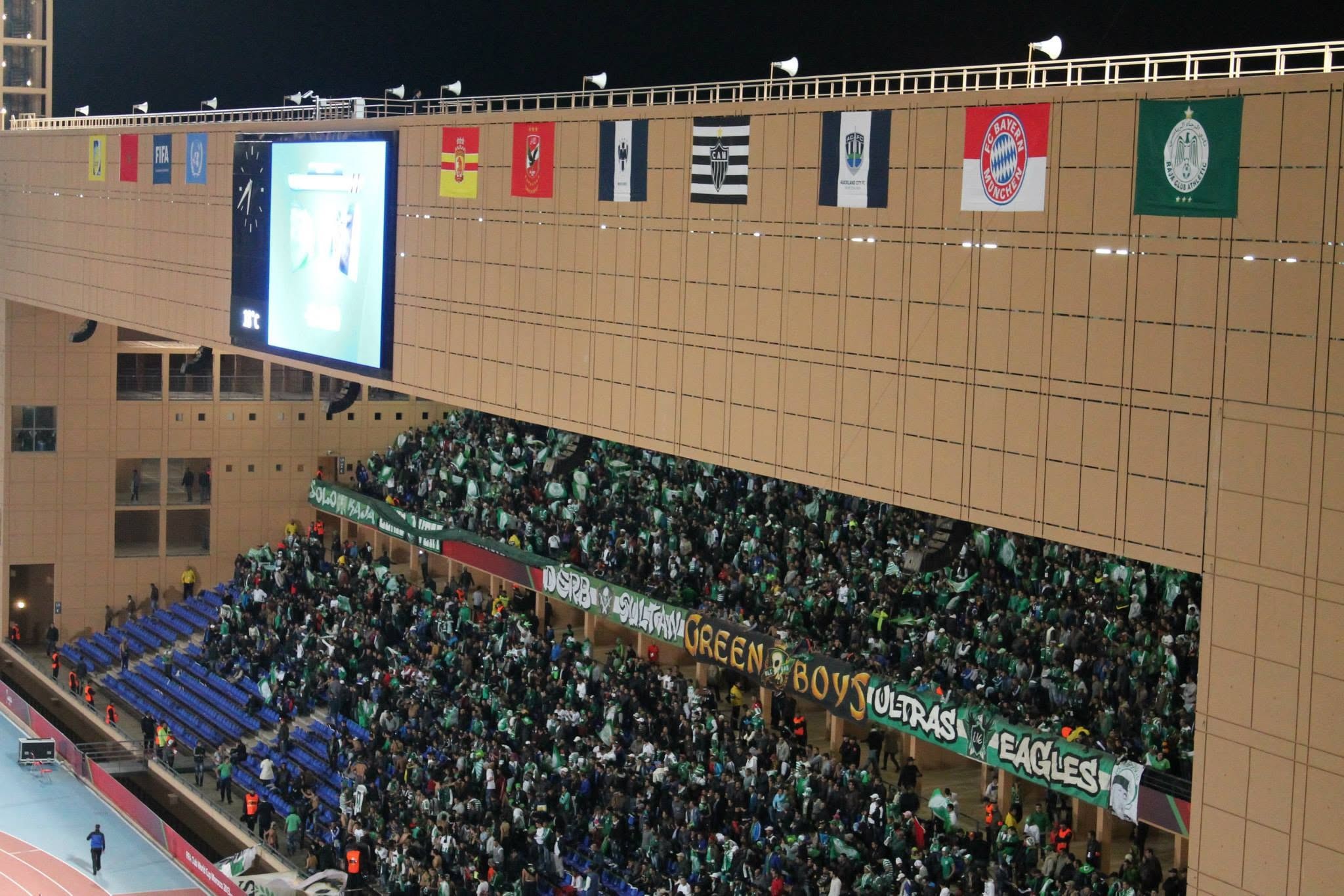 Vista a&eacute;rea de um est&aacute;dio de futebol lotado, com a maioria dos torcedores vestindo verde. Um tel&atilde;o exibe uma imagem clara de um trof&eacute;u brilhante. Acima do tel&atilde;o, diversas bandeiras de clubes e pa&iacute;ses adornam a estrutura do est&aacute;dio. Na parte inferior, uma pista de atletismo azul e vermelha &eacute; vis&iacute;vel, com uma pessoa caminhando. Faixas de torcidas organizadas como GREEN BOYS ULTRAS e EAGLES s&atilde;o exibidas nas arquibancadas
