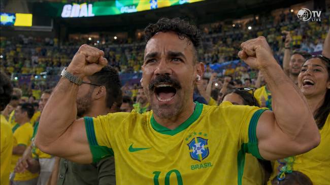 Homem de bigode e barba, vestindo camisa amarela da sele&ccedil;&atilde;o brasileira, comemora um gol com os bra&ccedil;os erguidos e punhos cerrados, boca aberta em grito de alegria. Ele est&aacute; em um est&aacute;dio lotado, com outros torcedores ao fundo, tamb&eacute;m celebrando. No placar, a palavra GOAL &eacute; vis&iacute;vel