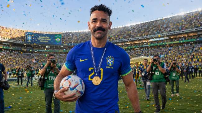 Homem de bigode e cavanhaque, vestindo camisa azul da sele&ccedil;&atilde;o brasileira com medalha dourada no pesco&ccedil;o, segura uma bola de futebol. Ele est&aacute; em um campo de futebol, com confetes coloridos caindo, e uma multid&atilde;o de torcedores nas arquibancadas ao fundo.