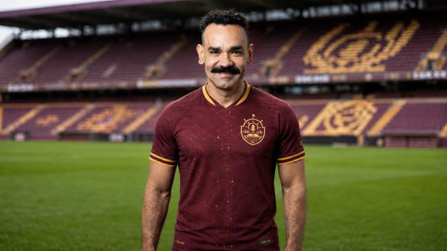 Imagem de IA mostra homem de pele morena, bigode e cabelo curto encaracolado, vestindo camisa de futebol vinho com detalhes dourados, em p&eacute; no gramado de um est&aacute;dio com arquibancadas marrons ao fundo