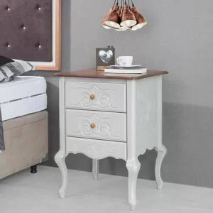 Mesa de cabeceira branca estilo proven&ccedil;al com tr&ecirc;s gavetas entalhadas e puxadores dourados, tampo marrom. Sobre ela, um porta-retrato, livros e uma x&iacute;cara. Ao fundo, uma cama e um lustre de cobre.