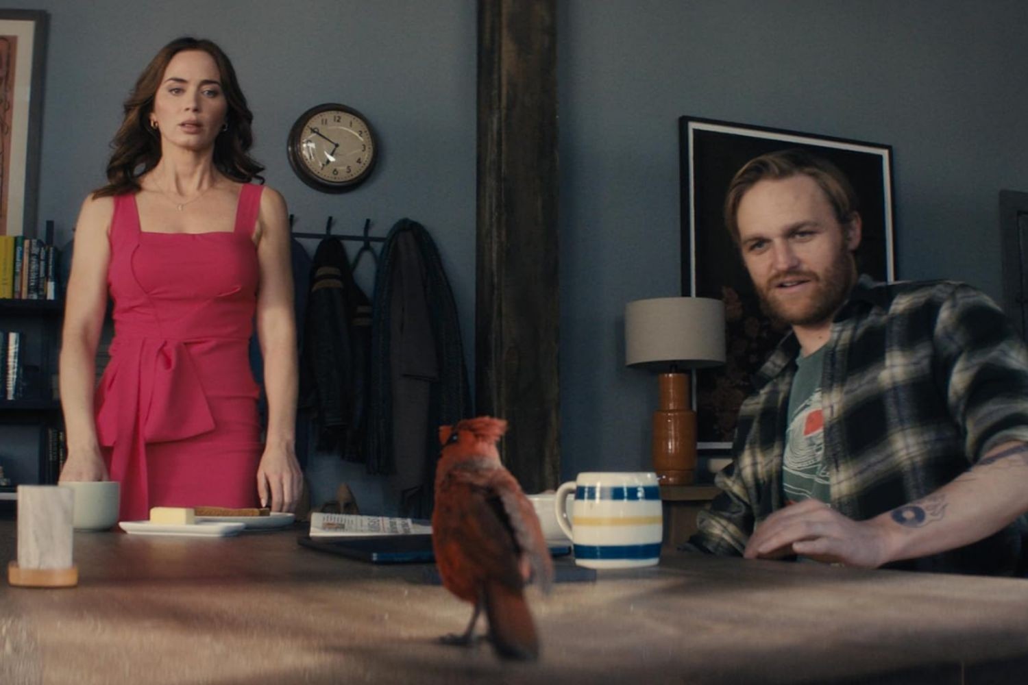 Uma mulher de vestido rosa choque e um homem barbudo de camisa xadrez sentados &agrave; mesa, observando um p&aacute;ssaro vermelho sobre ela. H&aacute; x&iacute;caras de caf&eacute; e um peda&ccedil;o de manteiga na mesa. Ao fundo, um rel&oacute;gio de parede e um quadro escuro.