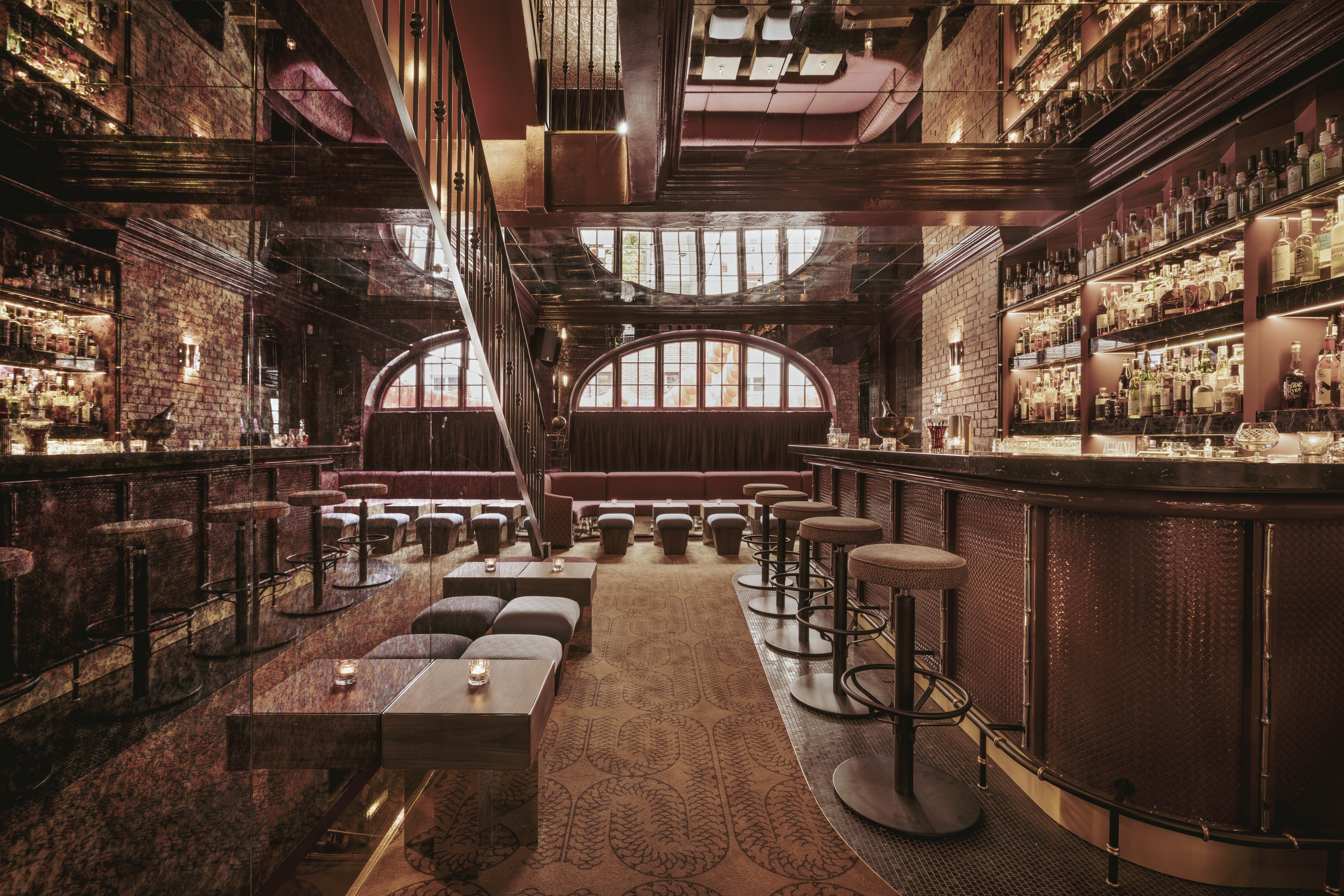 Interior de um bar sofisticado com ilumina&ccedil;&atilde;o baixa e tons terrosos. &Agrave; direita, um balc&atilde;o longo com banquetas estofadas e prateleiras cheias de garrafas. Ao fundo, janelas arqueadas e sof&aacute;s vermelhos. Uma escada escura sobe &agrave; esquerda, e mesas baixas com velas est&atilde;o no centro, sobre um tapete estampado. O teto espelhado reflete o ambiente, criando uma atmosfera aconchegante e elegante