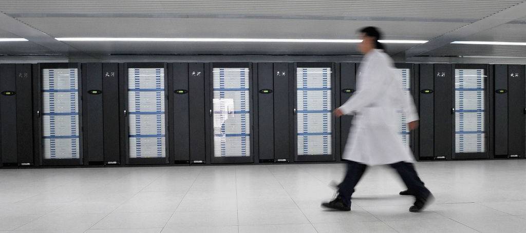 Pessoa de jaleco branco e calça escura caminhando rapidamente da direita para a esquerda em um corredor com fileiras de servidores de computador pretos e cinzas ao fundo