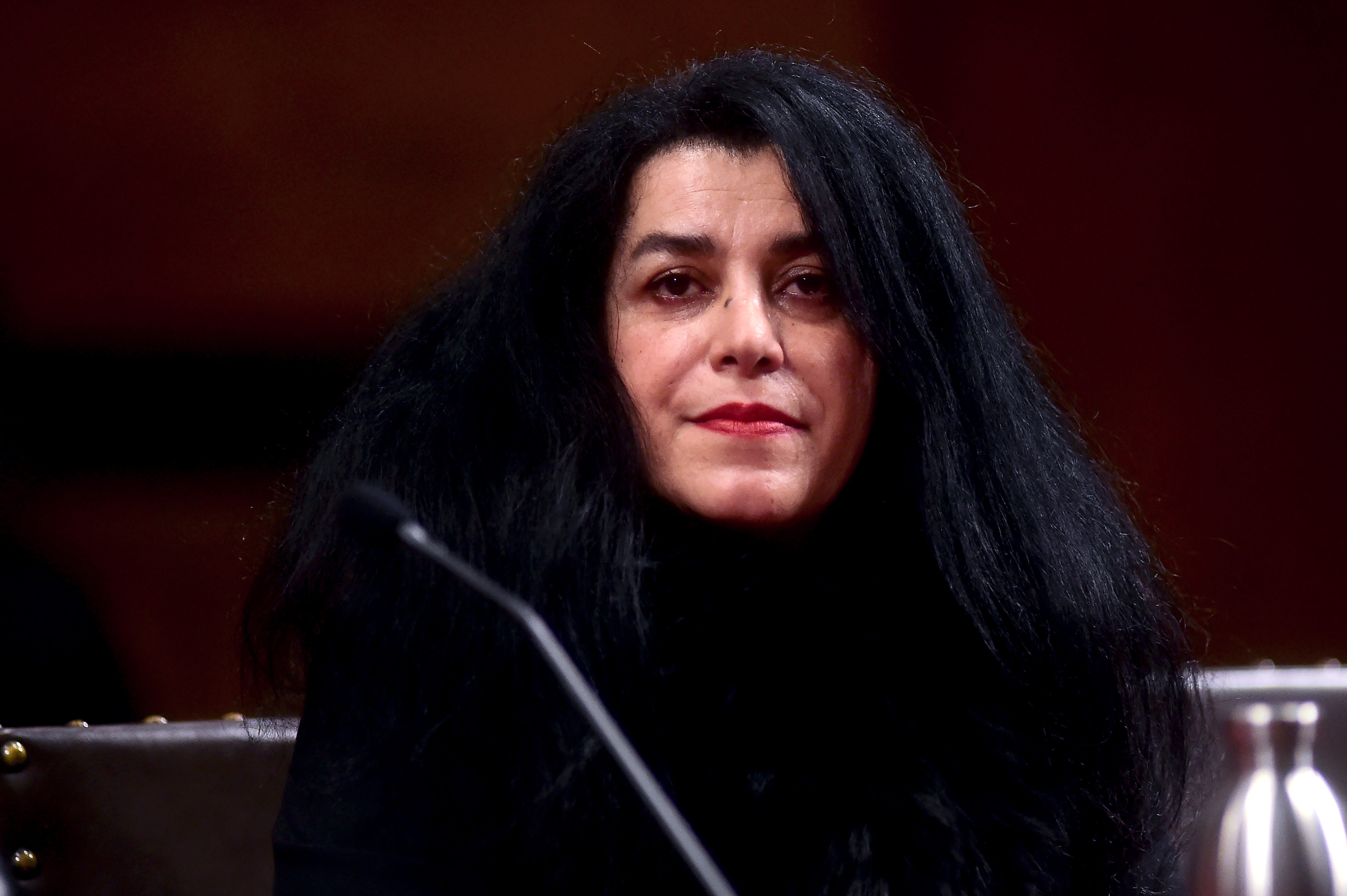 Mulher de pele clara e cabelos pretos longos, soltos e volumosos, com batom vermelho, olhando para frente. Ela veste roupa escura e est&aacute; sentada em um ambiente interno com fundo marrom avermelhado e um microfone &agrave; frente