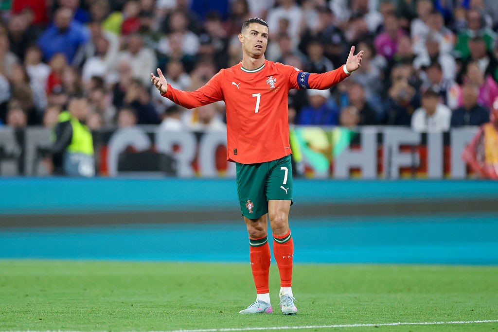 Cristiano Ronaldo, jogador de futebol, em campo com uniforme vermelho e verde, n&uacute;mero 7, bra&ccedil;os abertos em gesto de questionamento, em frente a uma plateia desfocada