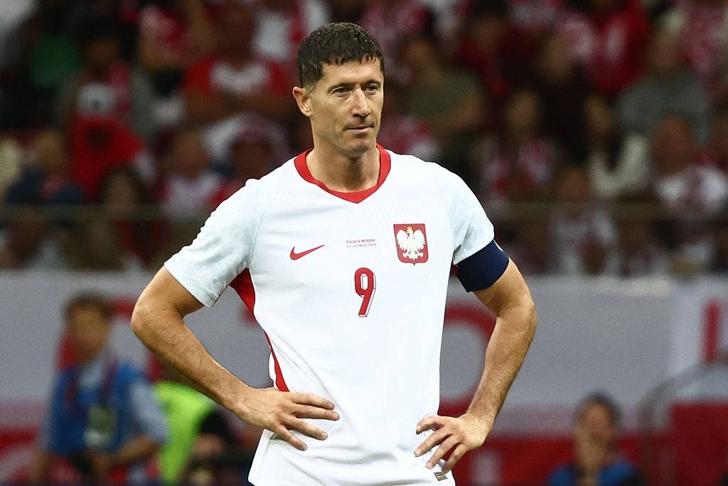 O jogador de futebol Robert Lewandowski, com cabelo curto e escuro, veste a camisa branca da seleção polonesa com detalhes vermelhos, o número 9 e o escudo da águia branca. Ele está em campo, com as mãos na cintura e expressão séria, olhando para o lado direito, com a torcida desfocada ao fundo