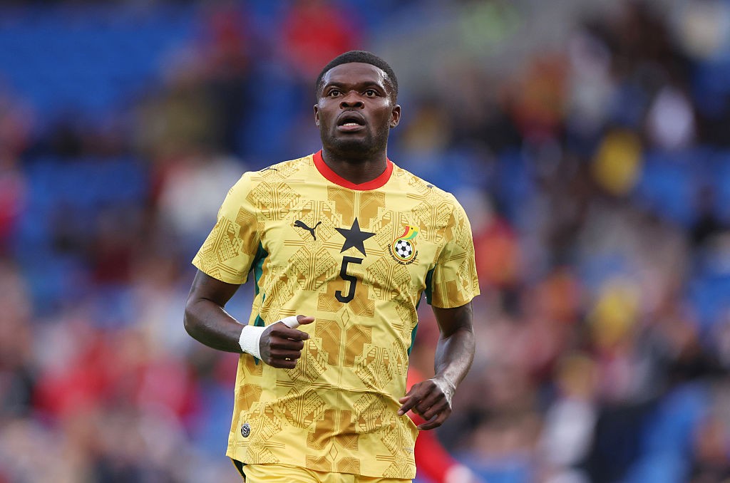 Jogador de futebol de Gana, pele escura, camisa amarela com detalhes em dourado, n&uacute;mero 5 e estrela preta no peito, olhando para cima com express&atilde;o s&eacute;ria, em campo de futebol com torcida desfocada ao fundo
