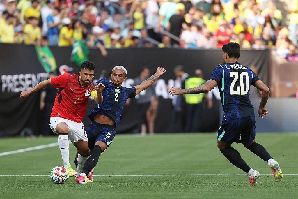 Três jogadores de futebol em campo. Um de camisa vermelha e shorts brancos tenta dominar a bola, enquanto outro de camisa azul escura e shorts azuis o marca de perto. Um terceiro jogador de camisa azul escura com o número 20 nas costas corre para a direita. Torcedores assistem ao fundo