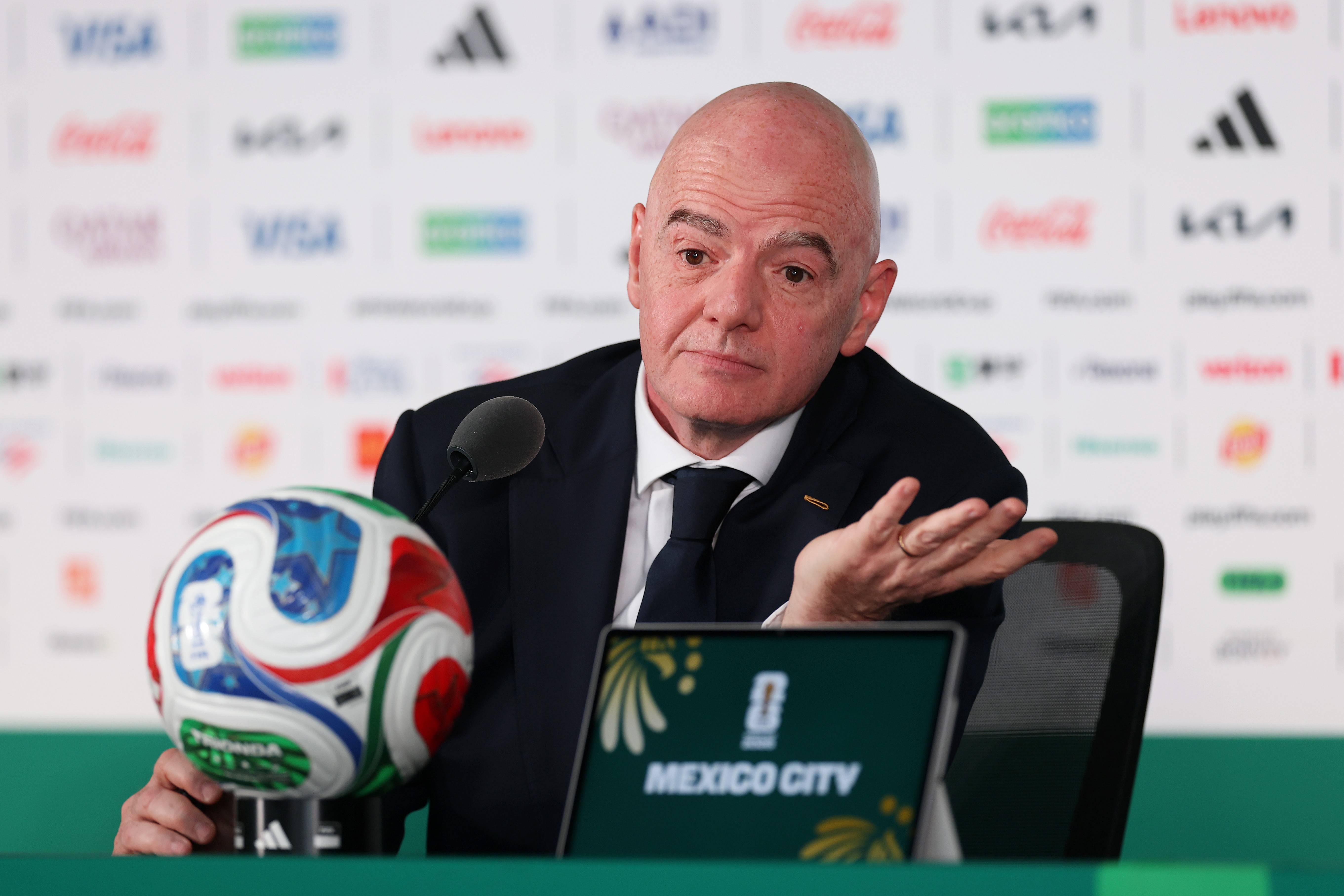 Gianni Infantino, presidente da FIFA, em coletiva de imprensa, gesticula com a mão direita. Ele veste terno azul-marinho, camisa branca e gravata escura. À esquerda, uma bola de futebol colorida. À frente, um tablet com MEXICO CITY em destaque. O fundo branco exibe logotipos de patrocinadores desfocados