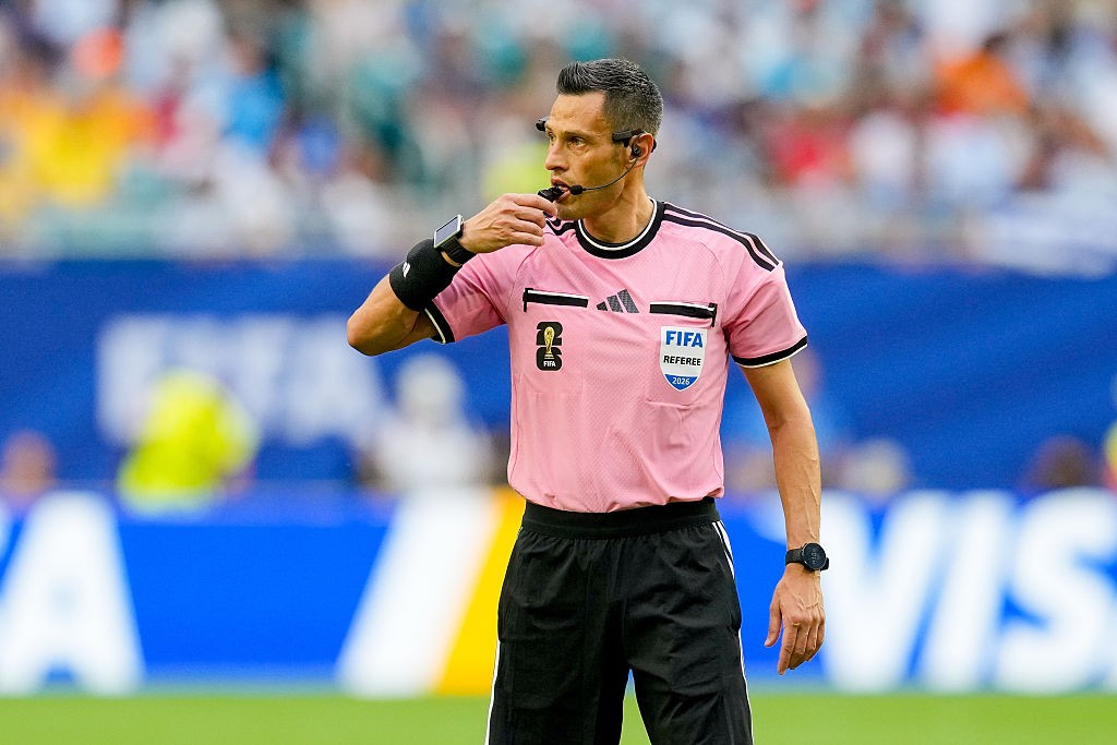 &Aacute;rbitro de futebol masculino, com cabelo grisalho e uniforme rosa, apitando em campo. Ele usa um rel&oacute;gio inteligente no pulso esquerdo e um fone de ouvido.