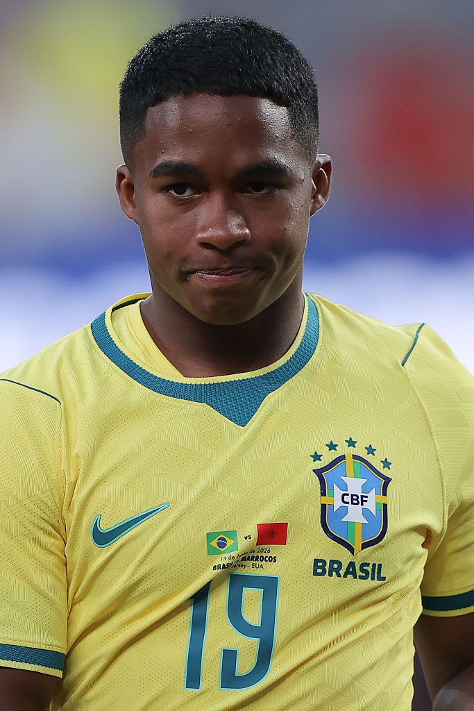 Jovem jogador de futebol negro, com cabelo curto e escuro, vestindo a camisa amarela da sele&ccedil;&atilde;o brasileira com gola azul e o n&uacute;mero 19 em destaque. Ele olha para frente com express&atilde;o s&eacute;ria, o escudo da CBF e a palavra BRASIL vis&iacute;veis na camisa.