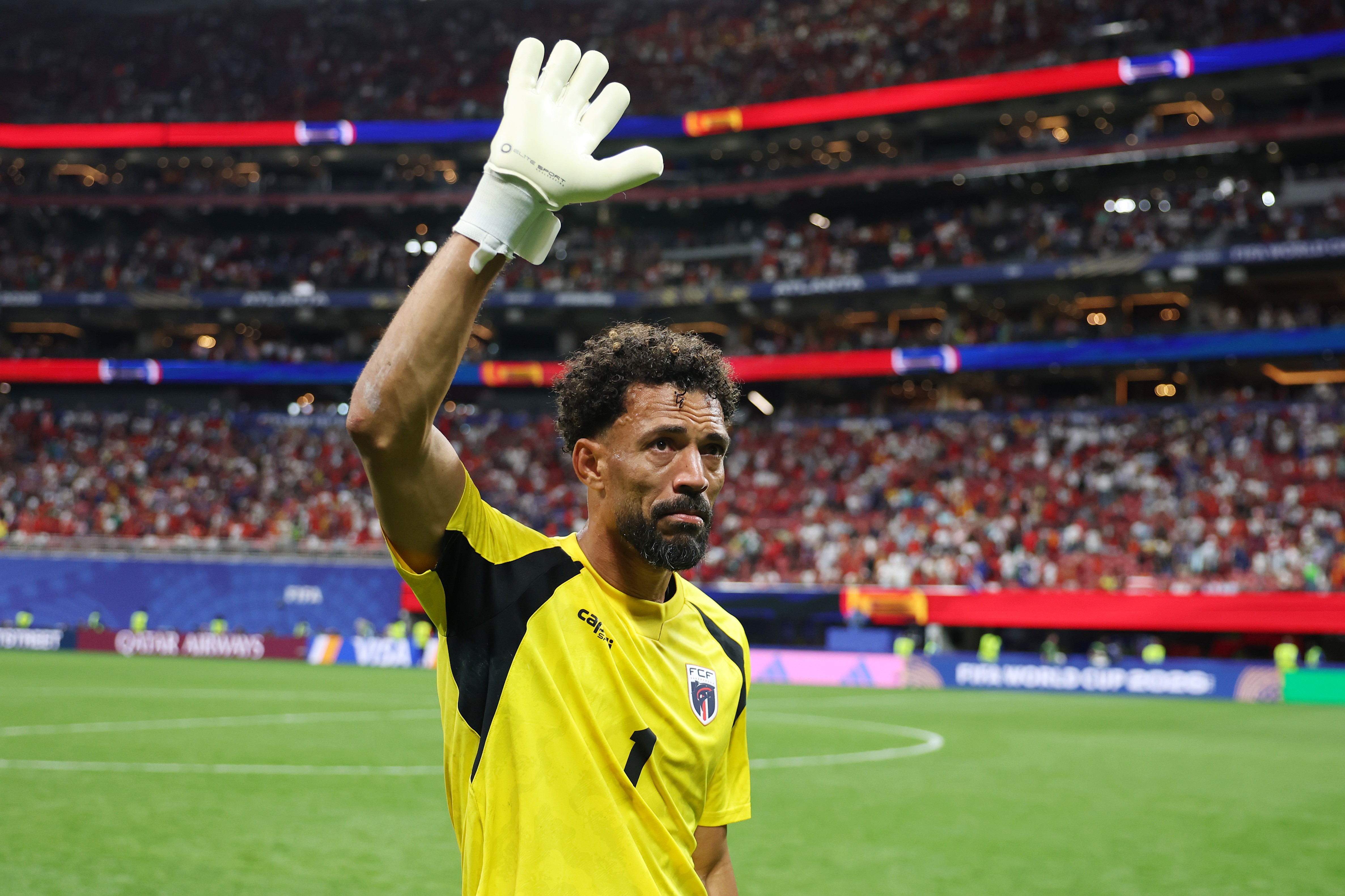 Goleiro de pele escura, barba e cabelo crespo, vestindo camisa amarela e luvas brancas, acena com a m&atilde;o direita para a torcida em um est&aacute;dio de futebol lotado