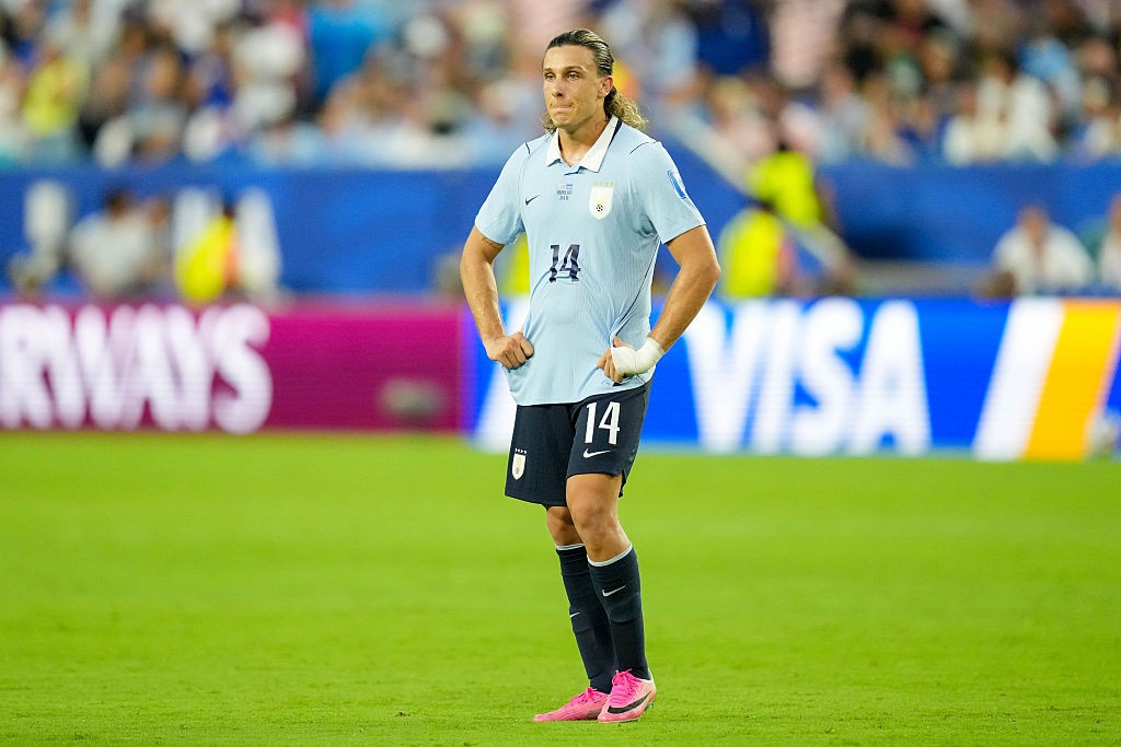 Jogador de futebol com camisa azul-clara e shorts pretos, n&uacute;mero 14, em p&eacute; no gramado verde de um est&aacute;dio, m&atilde;os na cintura, olhando para frente. Ele tem cabelos longos e loiros presos para tr&aacute;s, e uma faixa branca no punho esquerdo. Ao fundo, placas de publicidade com AIRWAYS e VISA e torcedores borrados.
