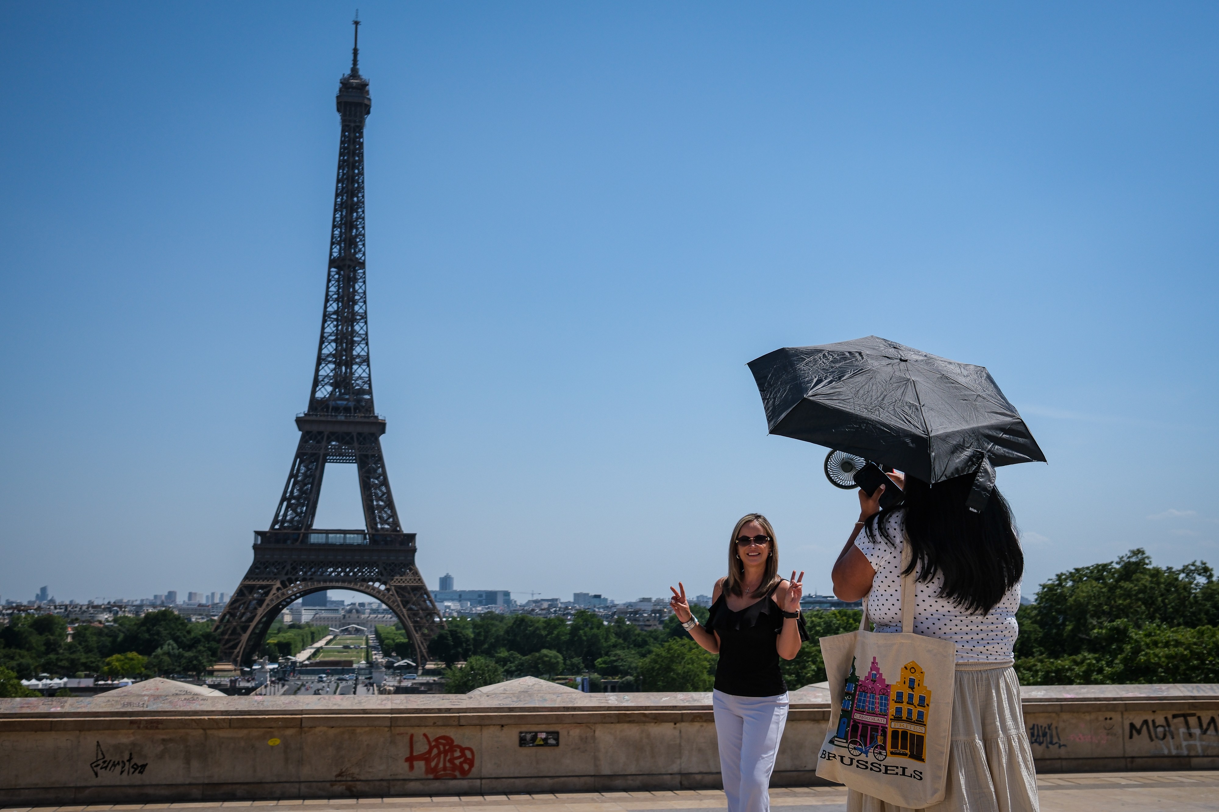 Duas mulheres em Paris. Uma loira de óculos escuros, blusa preta e calça branca faz sinal de paz. A outra, de costas, usa chapéu e segura um guarda-chuva preto, com uma bolsa de tecido com a estampa Brussels e casas coloridas. Ao fundo, a Torre Eiffel e a cidade sob céu azul