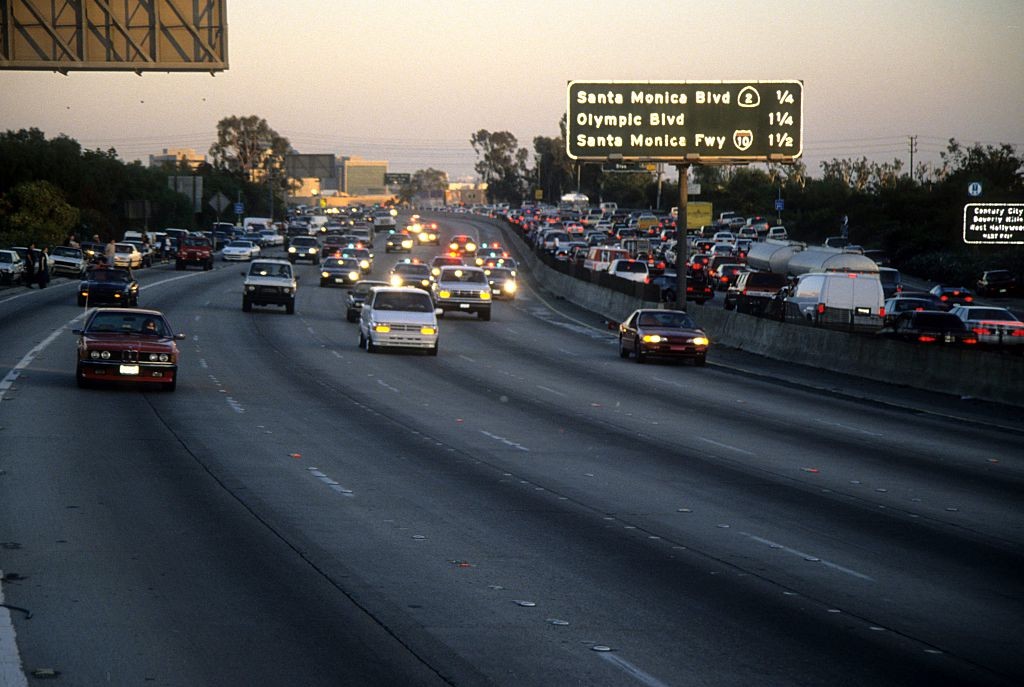 Rodovia movimentada ao entardecer, com v&aacute;rios carros em tr&ecirc;s pistas, alguns com far&oacute;is acesos. Um letreiro verde indica Santa Monica Blvd e Olympic Blvd.