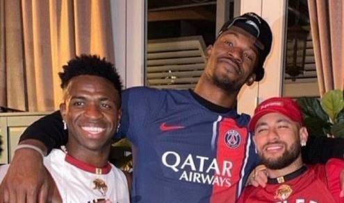 Três homens sorrindo para a câmera. Vinicius Jr. à esquerda, com camisa branca e colar vermelho, abraça Jimmy Butler, que usa boné preto e camisa do PSG. Neymar Jr. à direita, de boné e camisa vermelhos, sorri ao lado deles