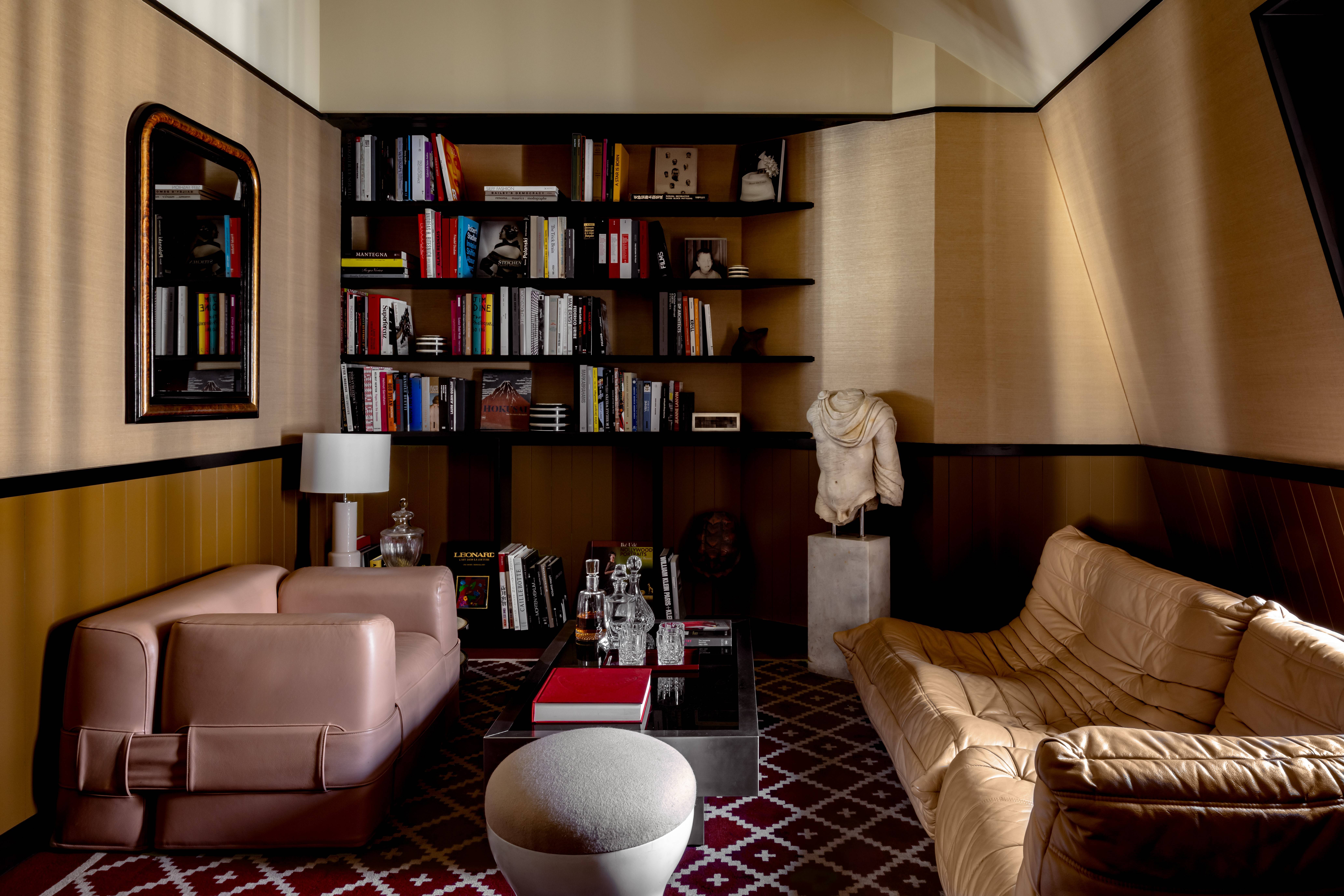 Sala de estar com paredes bege e marrom, estantes escuras repletas de livros coloridos, dois sofás de couro bege e uma mesa de centro com livros e garrafas. Um espelho retangular com moldura dourada reflete livros, e um busto de gesso branco está sobre um pedestal. O chão tem um tapete marrom com padrões geométricos brancos