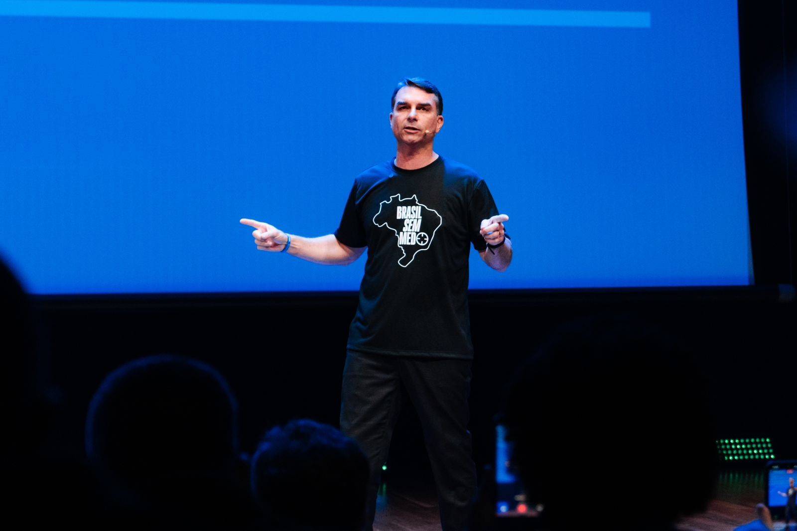Homem de meia-idade, cabelo escuro e camisa preta com estampa do mapa do Brasil e a frase Brasil Sem Medo, gesticula com os braços abertos e dedos apontados para os lados, em um palco escuro com um telão azul ao fundo