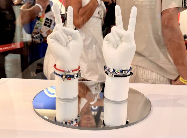 Duas m&atilde;os de manequim brancas, em formato de V de vit&oacute;ria, exibem pulseiras coloridas com pingentes tem&aacute;ticos sobre uma superf&iacute;cie espelhada. A m&atilde;o esquerda tem uma pulseira vermelha e outra azul com pingentes prateados e coloridos. A m&atilde;o direita exibe uma pulseira preta e outra azul com pingentes de esferas e um quadrado preto com a letra C. Pessoas desfocadas aparecem ao fundo