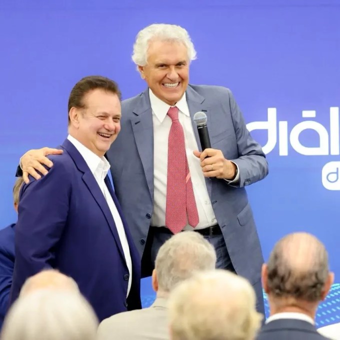 Dois homens sorridentes em um palco azul com a inscrição Diálogo da Sociedade. O da esquerda, de terno azul e camisa branca, é abraçado pelo da direita, de terno cinza, camisa branca e gravata vermelha, que segura um microfone. Pessoas assistem de costas em primeiro plano