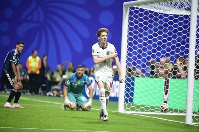 Jogador de futebol ingl&ecirc;s de cabelo ruivo, camisa branca e shorts, caminhando perto do gol com a bola dentro da rede. Um goleiro ajoelhado e outro jogador de camisa azul observam, em um campo verde com fundo azul