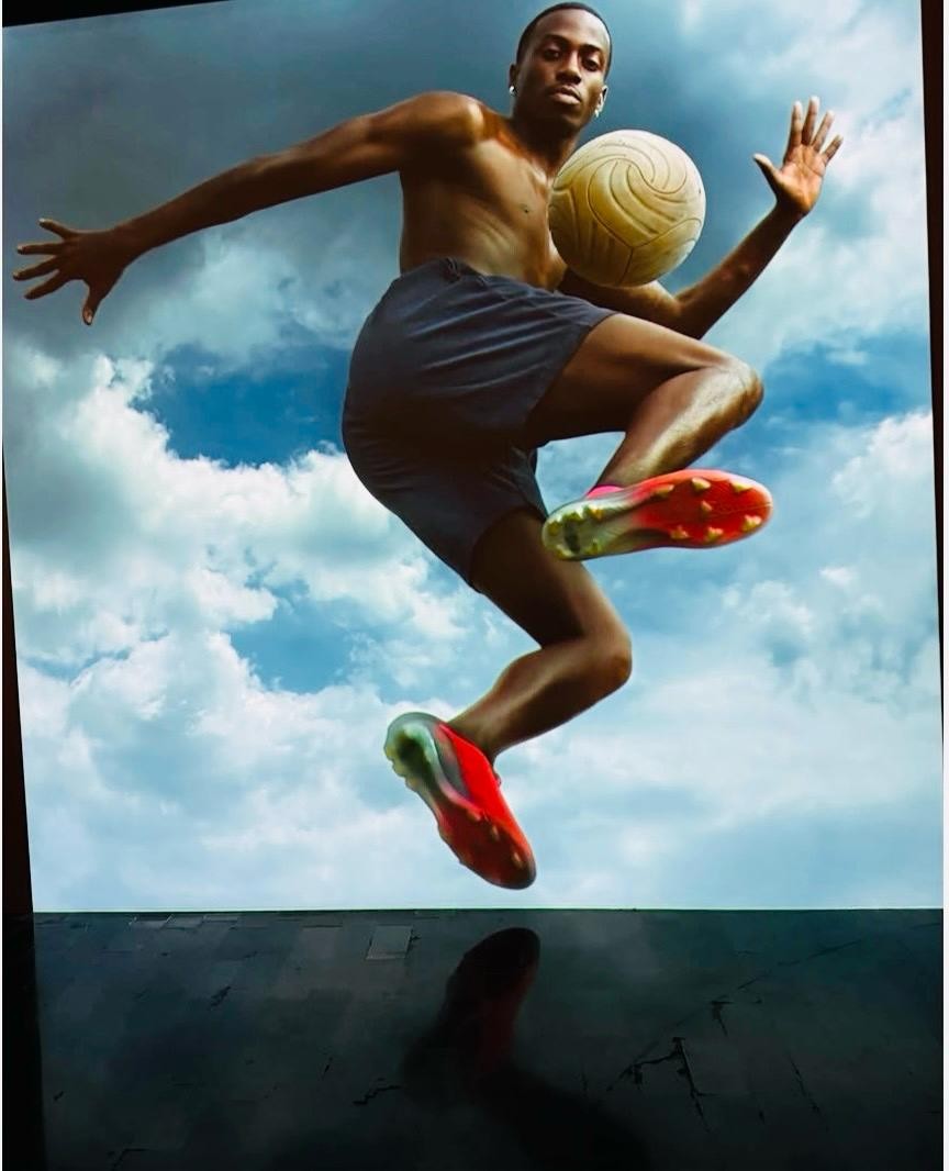 Homem negro sem camisa, usando shorts escuros e chuteiras vermelhas, salta no ar com uma bola de futebol amarela perto do peito, contra um c&eacute;u azul com nuvens brancas. Sua sombra &eacute; projetada em uma superf&iacute;cie escura abaixo.