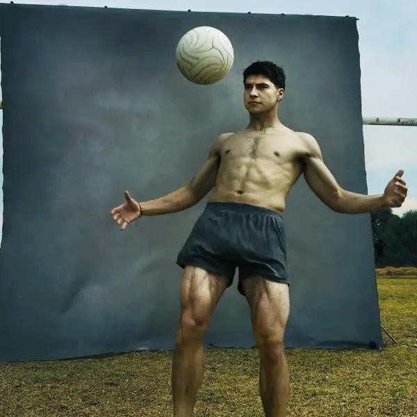 Homem sem camisa, com shorts escuros, equilibrando uma bola de futebol na cabe&ccedil;a, bra&ccedil;os abertos, em frente a um fundo cinza e gramado