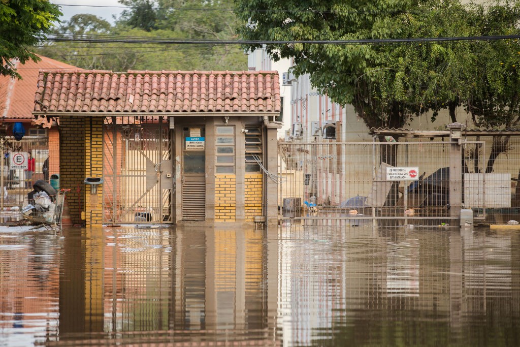 Rua alagada com &aacute;gua barrenta refletindo o c&eacute;u e as constru&ccedil;&otilde;es. &Agrave; esquerda, um port&atilde;o de tijolos e telhado de barro, com um carrinho de compras cheio de lixo. &Agrave; direita, um port&atilde;o de metal com uma placa de proibido estacionar. &Aacute;rvores e pr&eacute;dios ao fundo, sob um c&eacute;u nublado.