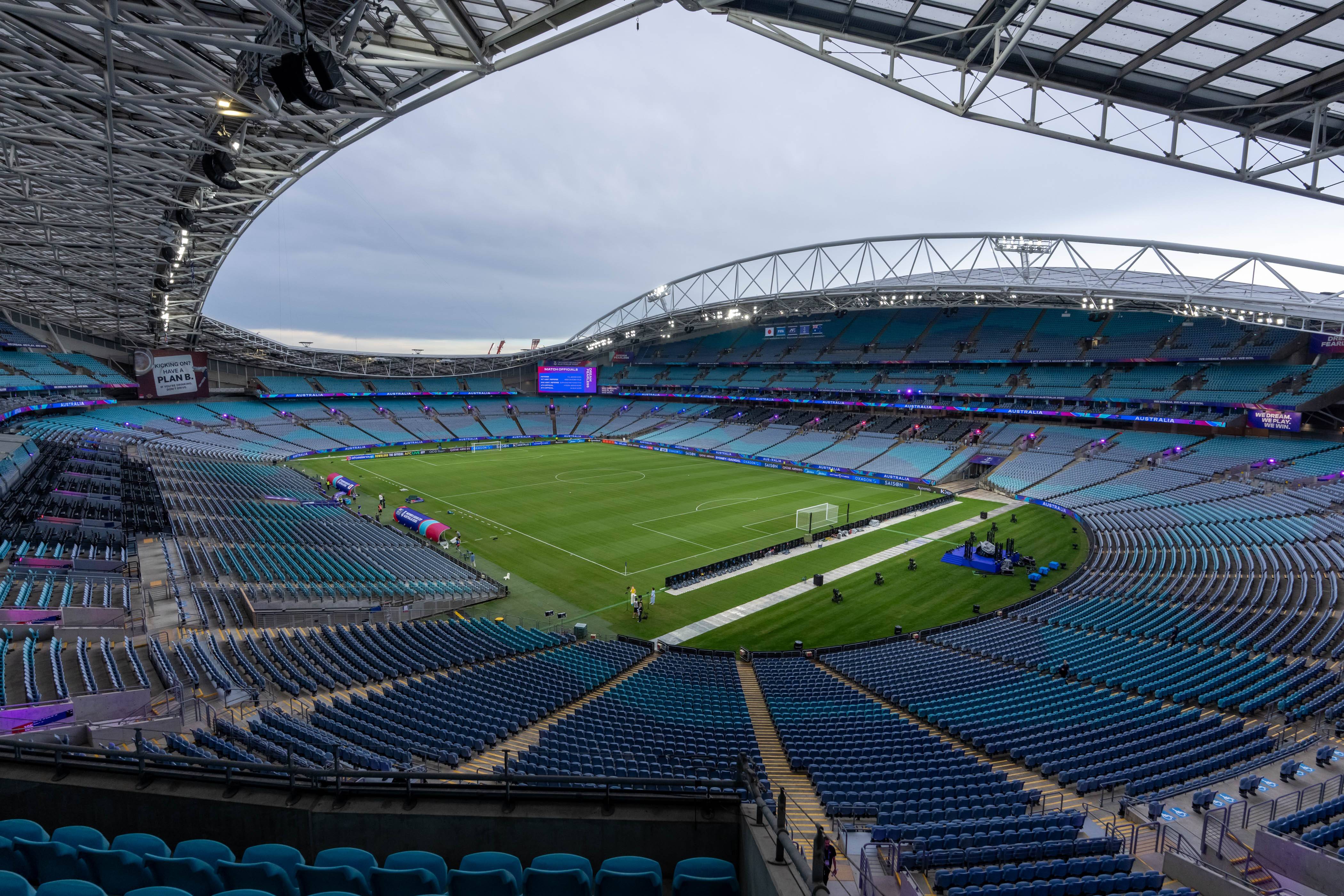 Vista panorâmica de um estádio de futebol moderno e vazio, com assentos azuis e verdes em camadas, gramado verde vibrante e um teto curvo de estrutura metálica. O céu está nublado