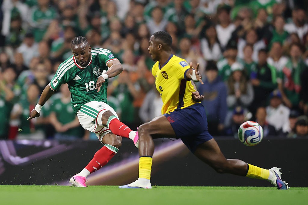 Dois jogadores de futebol, um de camisa verde e outro de camisa amarela, disputam a bola em campo verde. O jogador de camisa verde, com tran&ccedil;as, chuta a bola com a perna direita, usando chuteiras rosa. O jogador de camisa amarela, com cabelo curto, tenta interceptar a jogada. A bola est&aacute; no ar, entre eles. Torcedores desfocados em verde e branco aparecem ao fundo