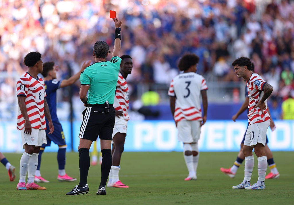 &Aacute;rbitro de futebol de costas, vestindo camisa verde e shorts pretos, levanta um cart&atilde;o vermelho. Jogadores com camisas listradas brancas e vermelhas, e um com camisa azul, observam no campo de grama verde, com torcida ao fundo