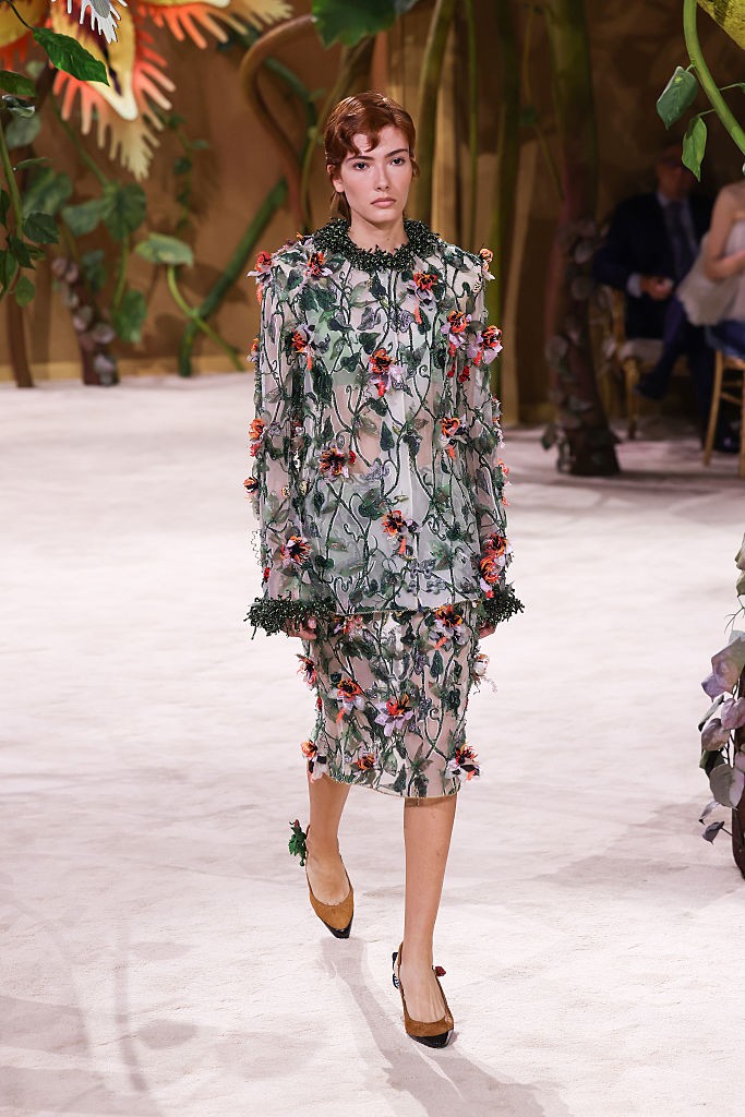 Modelo com cabelo ruivo curto, vestindo um conjunto transparente de saia e blusa com bordados de folhas verdes e flores coloridas, desfila em passarela clara com cen&aacute;rio de jardim bot&acirc;nico