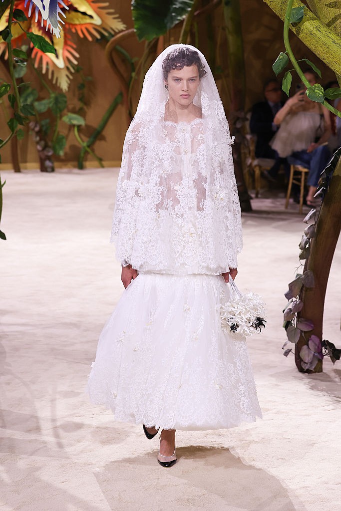 Modelo com cabelo cacheado escuro, usando um vestido de noiva branco rendado com v&eacute;u e um buqu&ecirc; de flores brancas, desfila em passarela clara com decora&ccedil;&atilde;o de folhagens e &aacute;rvores ao fundo